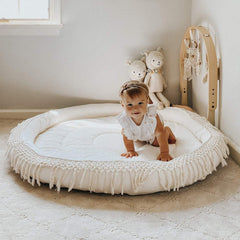 Baby standing on a round white mat in a room with toys and a window.