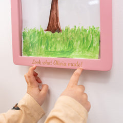 Child's hands pointing at a pink frame with a child's drawing of grass and a boot.