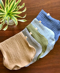 Set of five folded baby pants in beige, green, blue, and gray on a wooden surface with a plant in the corner.
