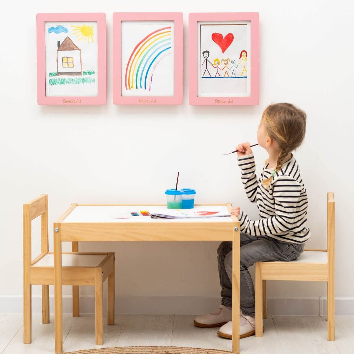 Child sitting at a wooden table with art supplies, drawing on paper, in a room with children's drawings on the wall.