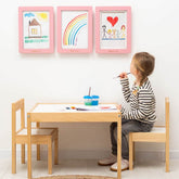 Child sitting at a wooden table with art supplies, drawing on paper, in a room with children's drawings on the wall.