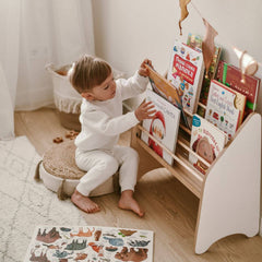 Child in a white outfit sitting on a stool, reaching for books on a wooden shelf.