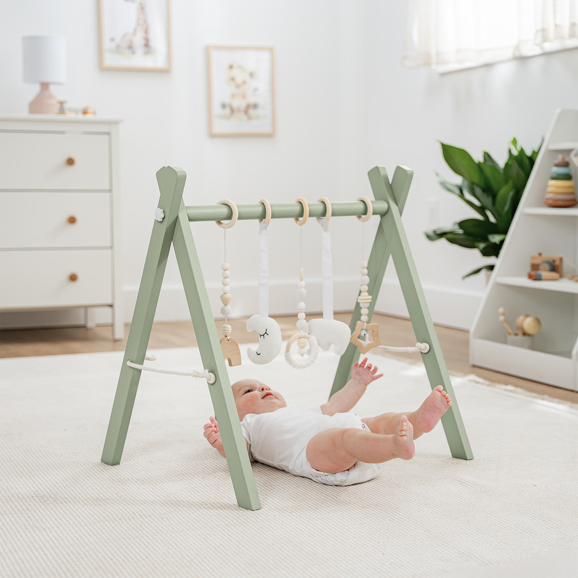 Baby lying on a white rug next to a green baby gym in a room with a dresser and plant.