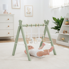 Baby lying on a white rug next to a green baby gym in a room with a dresser and plant.