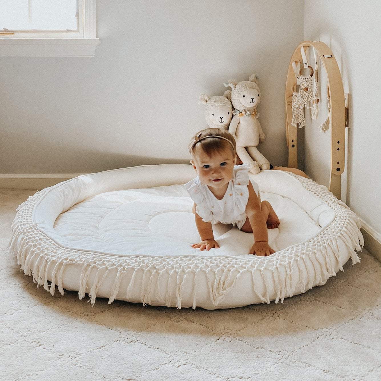 Baby standing on a round white mat in a room with toys and a window.