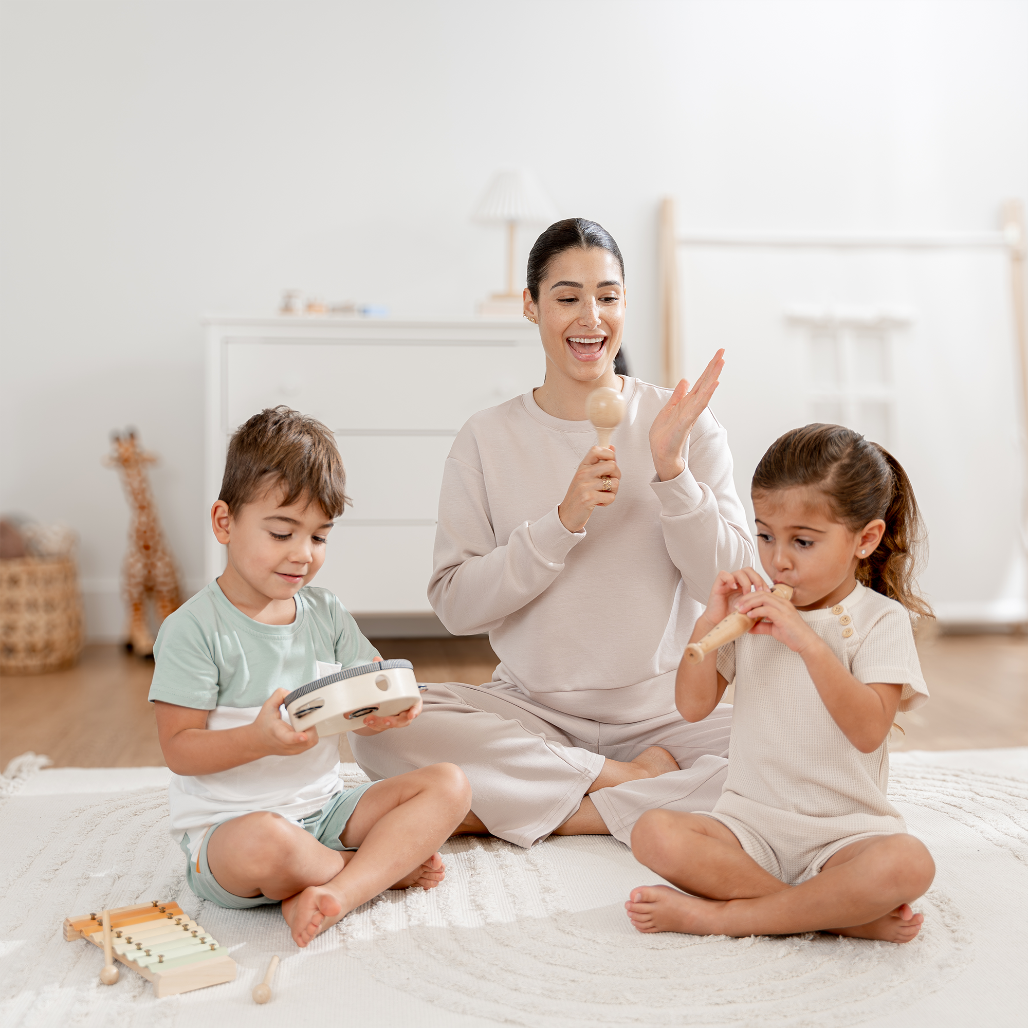 Woman and two children playing musical instruments on a bed in a bright room.