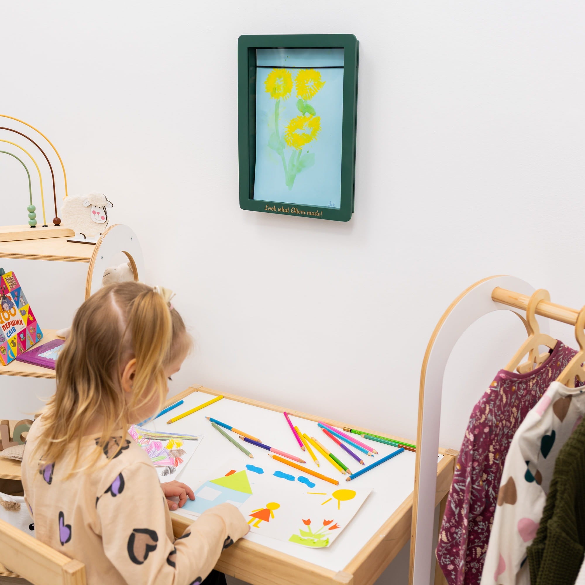 Child sitting at a table with art supplies, looking at a framed picture of flowers on the wall.