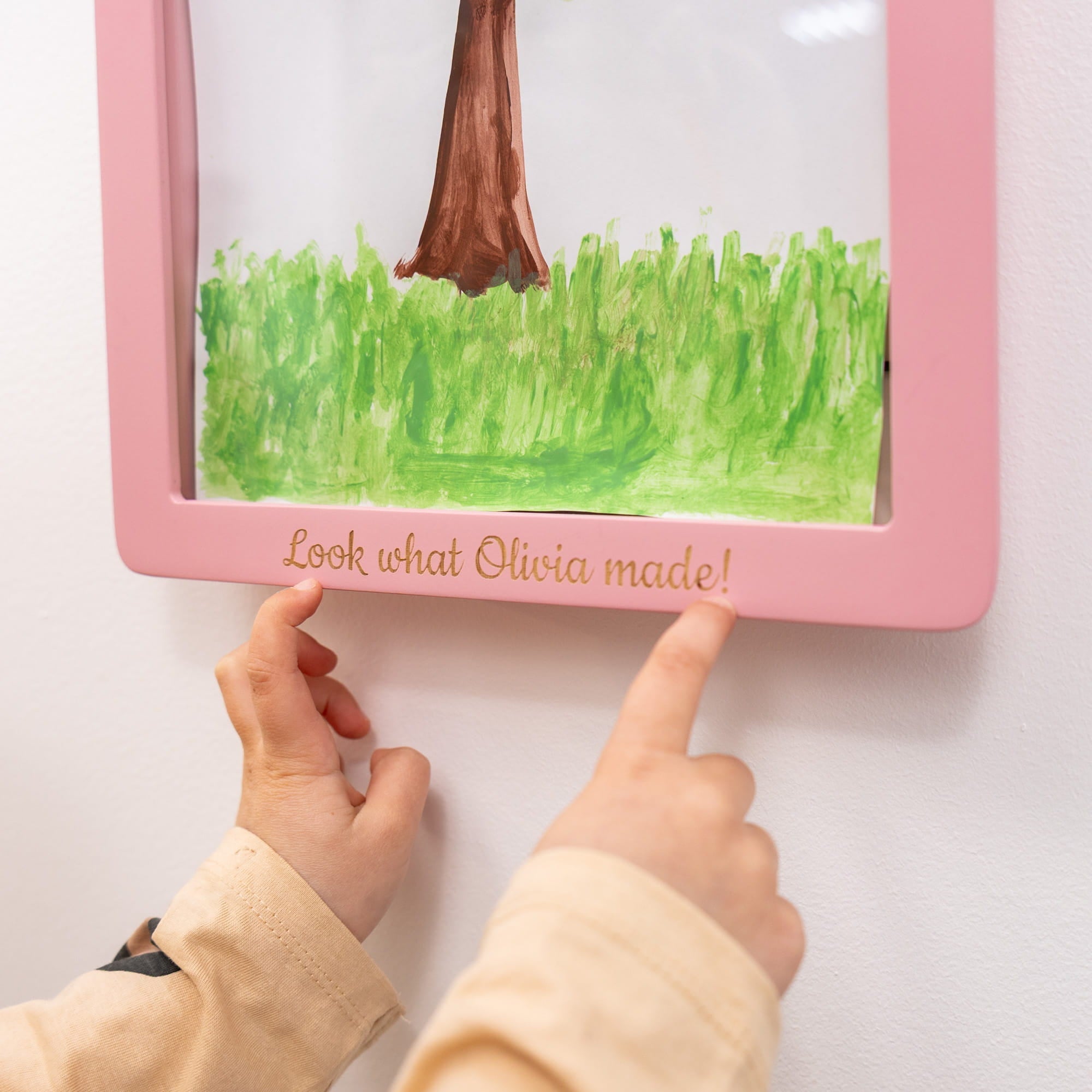 Child's hands pointing at a pink frame with a child's drawing of grass and a boot.