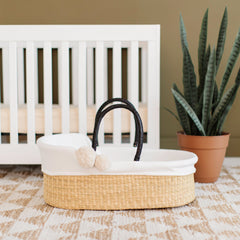 Wicker Moses basket with white cushion and black handle in front of a white crib with a potted plant.