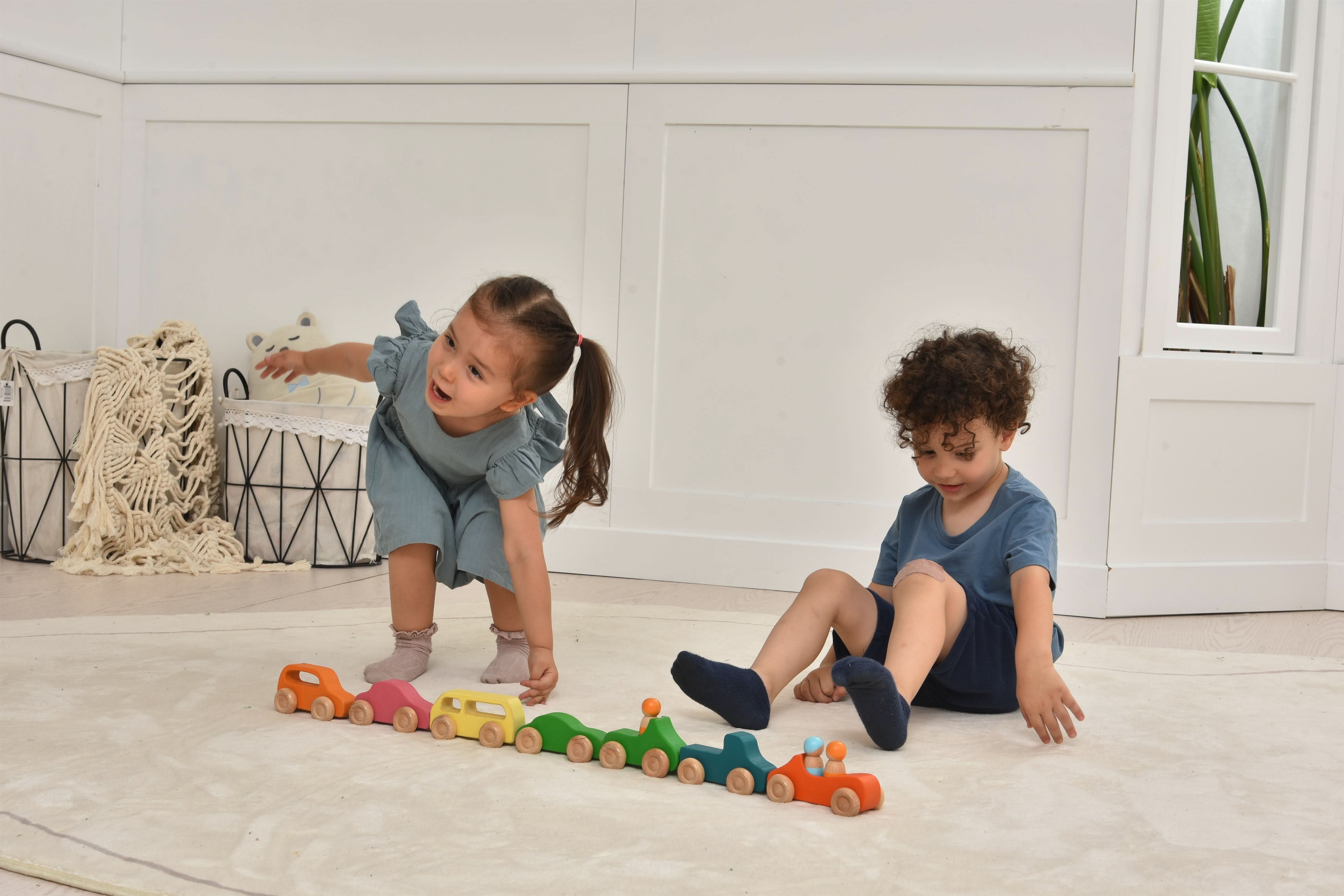 Two children playing with a set of colorful wooden toy cars on a light wood floor.