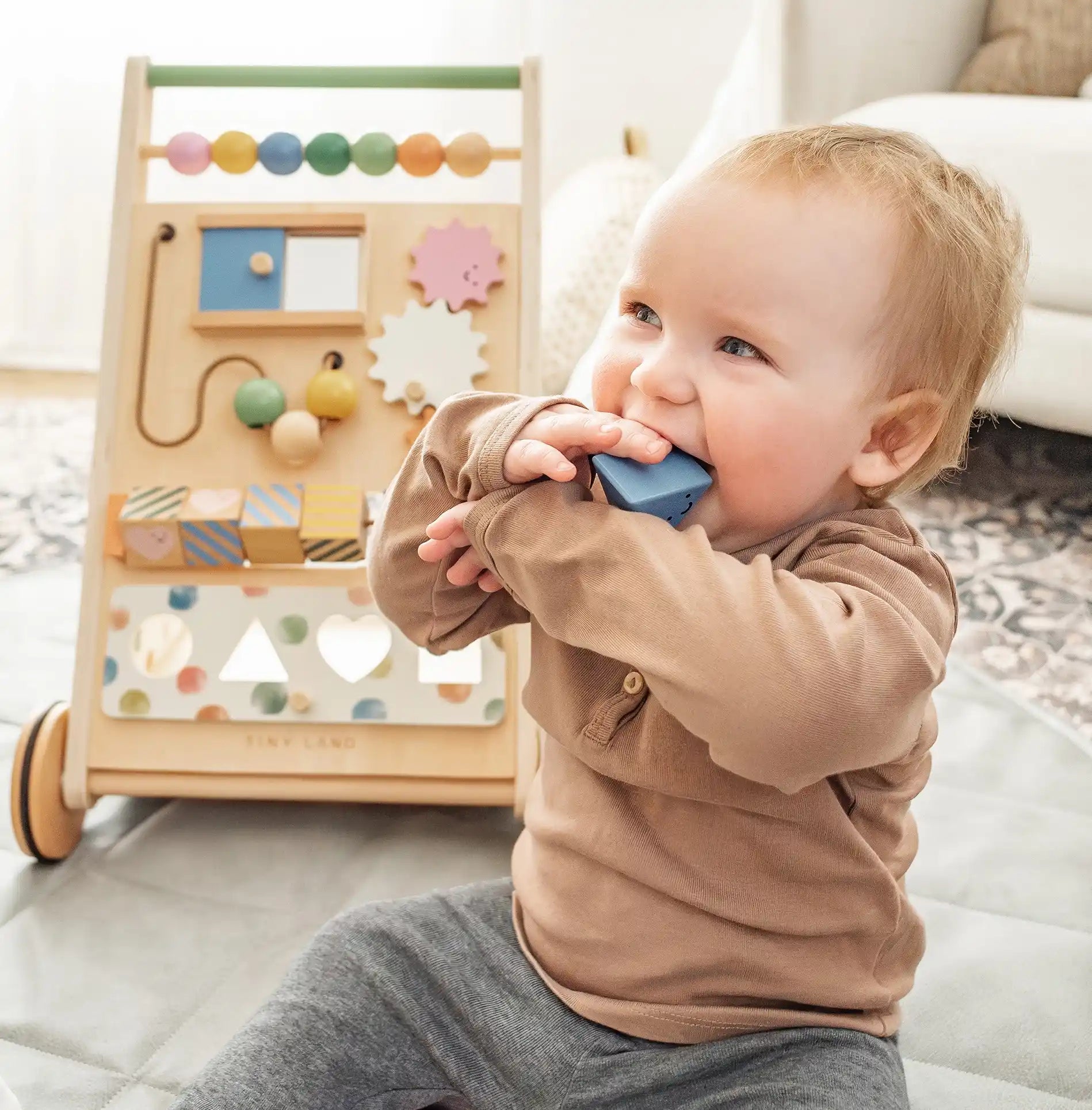 Child chewing on wooden block with activity walker in the background