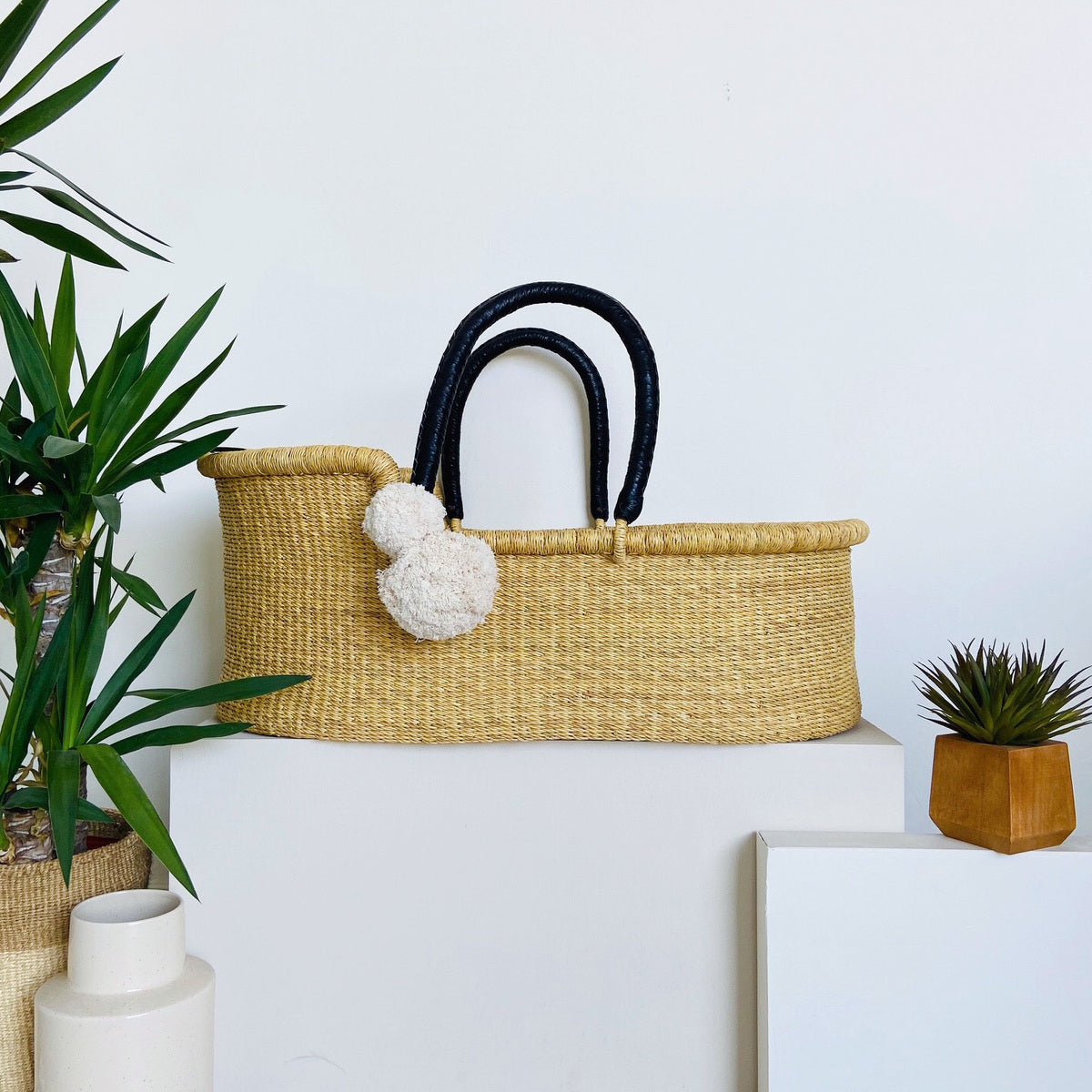 Woven basket with black handles on a white surface with plants around