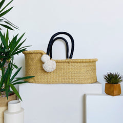 Woven basket with black handles on a white surface with plants around