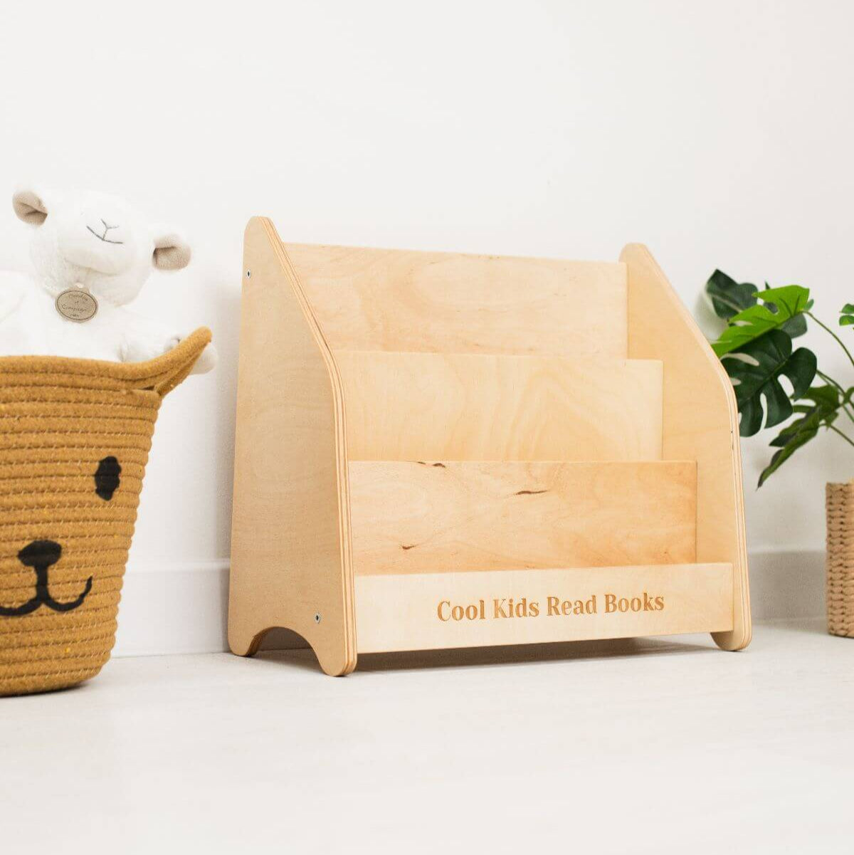 Wooden bookshelf with 'Cool Kids Read Books' text, next to a basket and plant on a white surface.