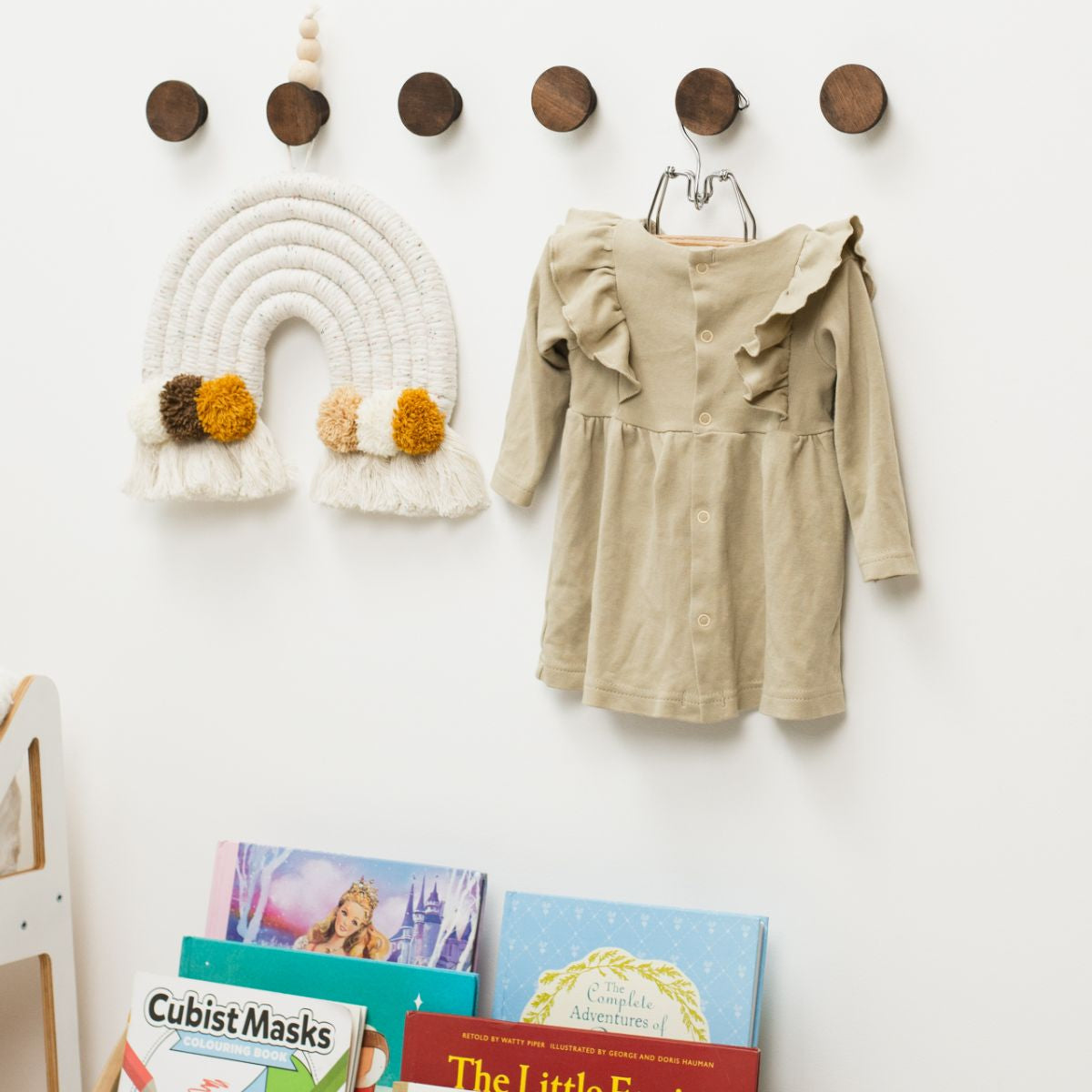 Children's dress hanging on a hook with books and decorative items below on a white wall.