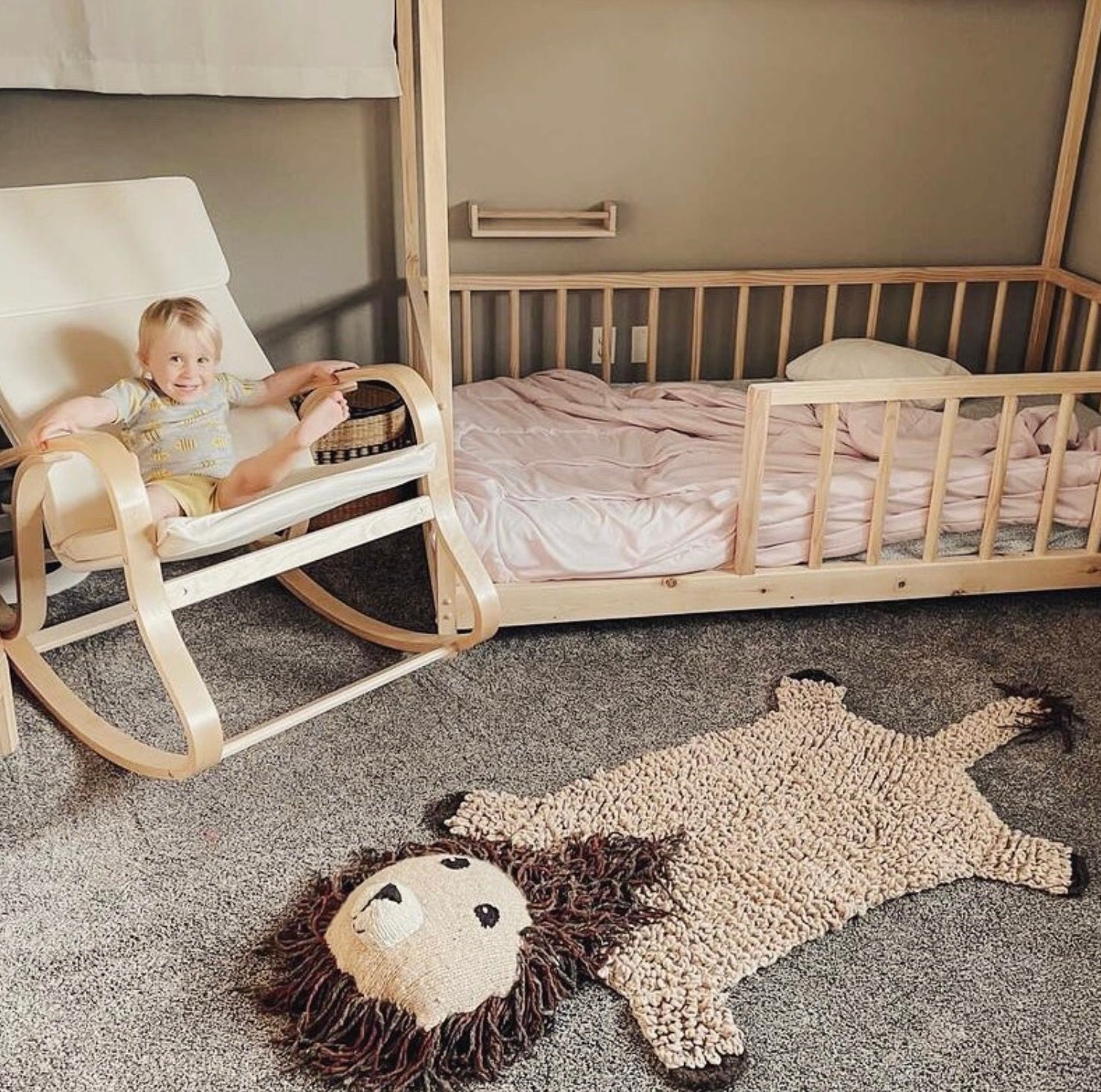 Child in a rocking chair next to a crib with a lion-shaped rug on the floor.