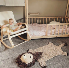 Child in a rocking chair next to a crib with a lion-shaped rug on the floor.
