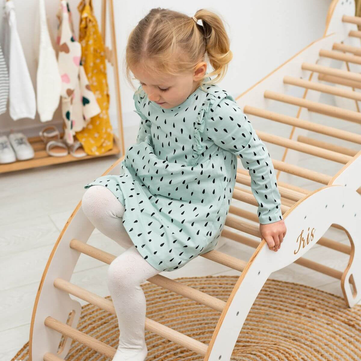 Child in a light green dress with black patterns sitting on a wooden chair.