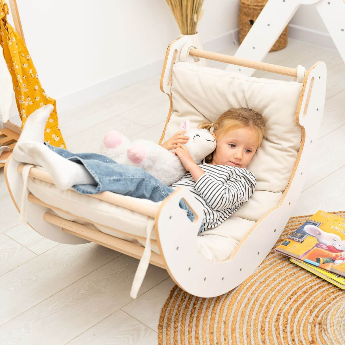 Child in a baby swing holding a plush toy in a cozy room.