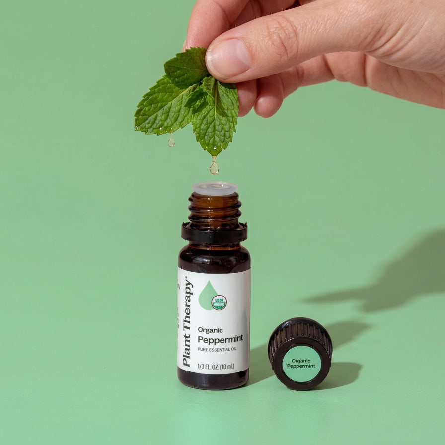 Bottle of Plant Therapy Organic Peppermint essential oil with a hand holding mint leaves on a green background