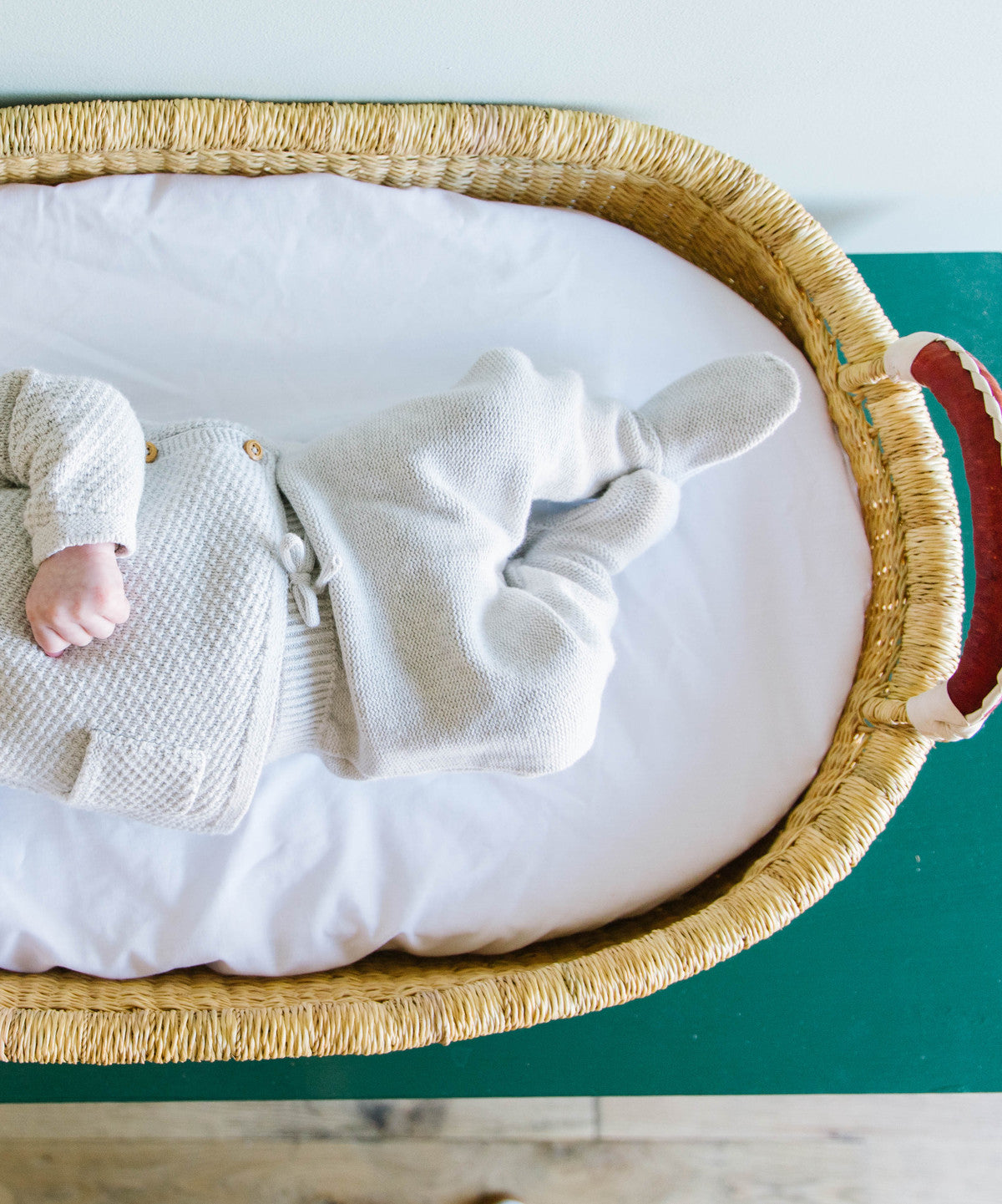 Baby in a white outfit lying on a white mattress inside a wicker crib with a green background.