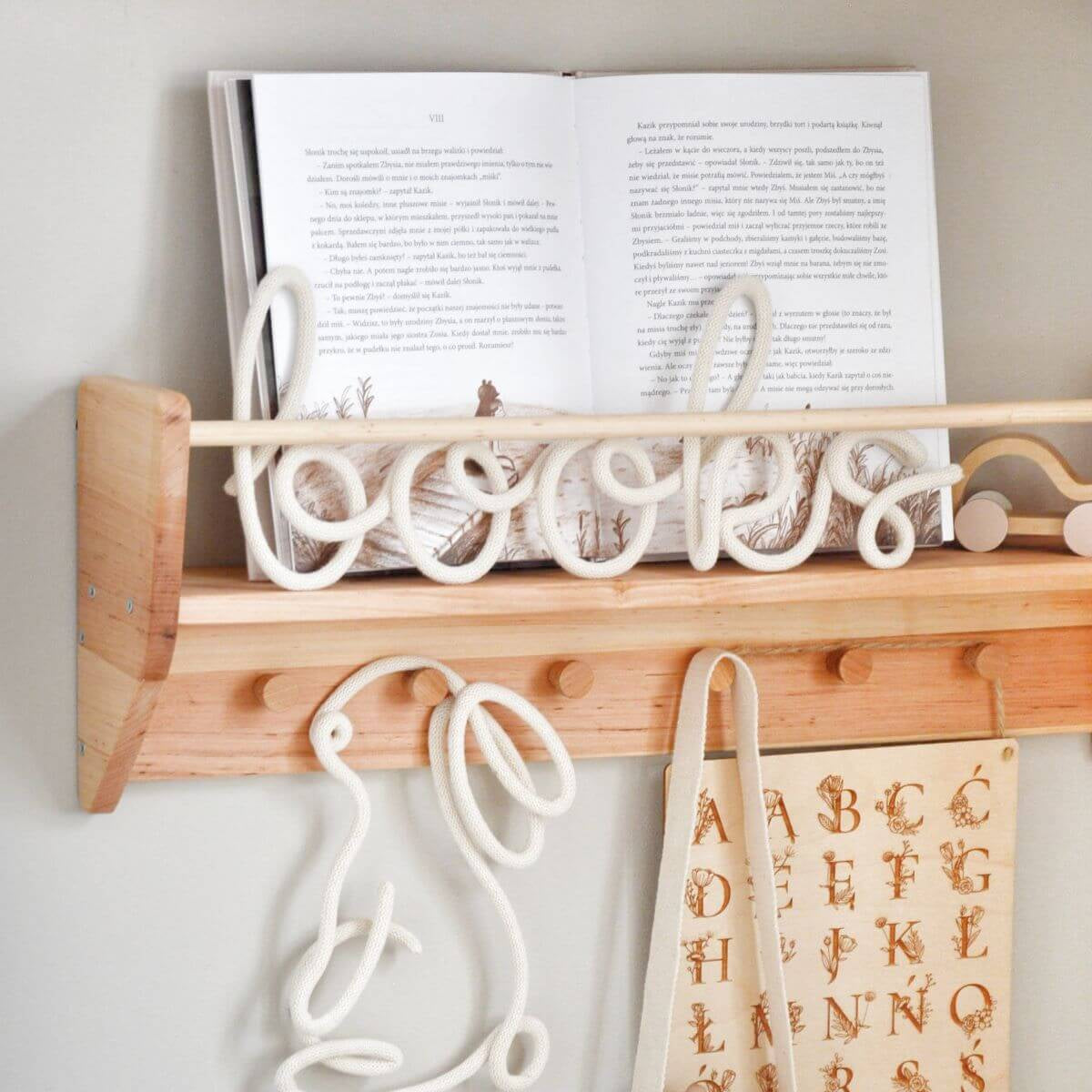 Wooden shelf with hooks holding an open book and a towel with letters on a light gray wall.