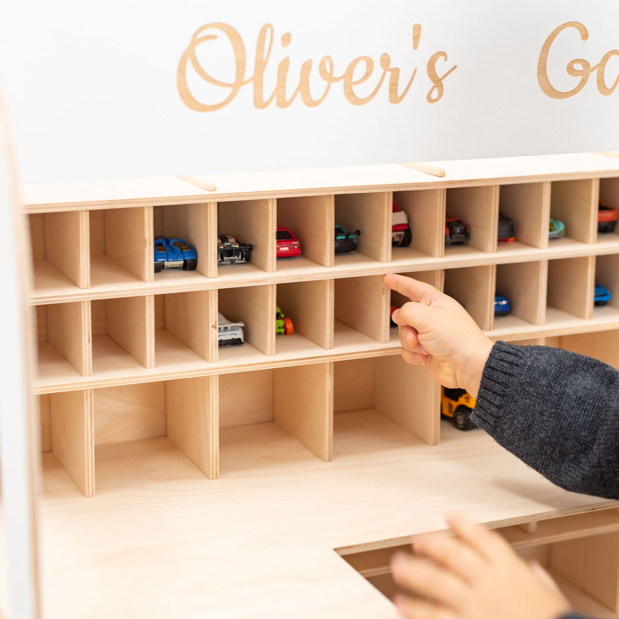 Wooden toy car organizer with small cars inside, labeled 'Oliver's', with a hand pointing at it.